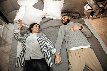 Pleasant family jumping. Beaming joyful couple connecting their hands while lying on bed in furniture storeの写真素材