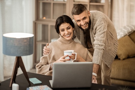 Guy hugging wife. Peaceful good-looking couple spending time together in cabinet while observing content in internetの写真素材