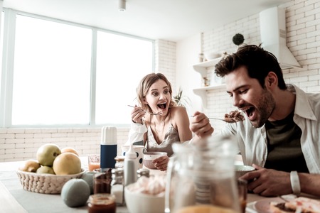 Joyful breakfast. Bearded handsome positive man in a white shirt feeling wonderful while eating having breakfast with his wifeの写真素材