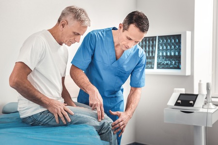 Blue uniform. Dark-haired experienced doctor wearing blue uniform examining knee of pensioner after his falling downの写真素材