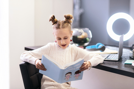 Funny book. Cute blue-eyed long-haired girl wearing bright earrings sitting in the chair while reading a funny bookの写真素材