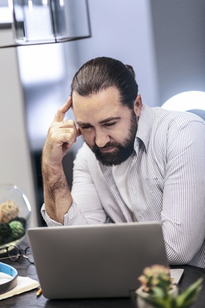 Thinking process. Dark-haired bearded man wearing a striped shirt looking serious