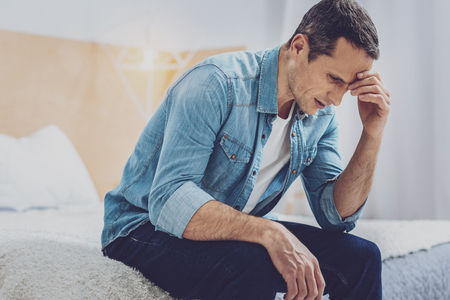 Bad news. Serious brunette male wrinkling forehead and bowing head while sitting at homeの写真素材