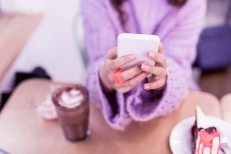 Chocolate cake nearby. Girl with accurate pink manicure typing messages on smartphone while being surrounded with goodiesの写真素材