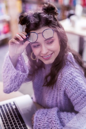 Working with laptop. Beaming fashionable lady with dark wavy hair taking of her clear glasses