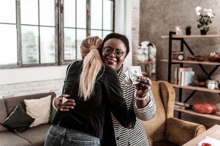 Friendly hugs. Young long-haired young woman wearing a black jacket holding hugging a dark-skinned...