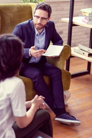 Having a session. Nice bearded man holding a notebook while having a conversation with his patientの写真素材