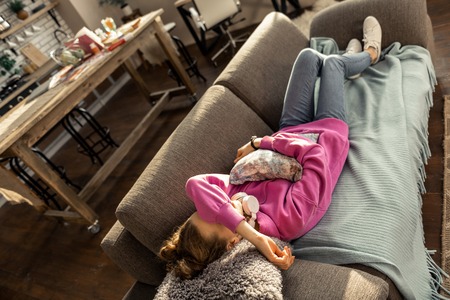 Lying on sofa. Slim teenage girl wearing jeans hugging little pillow and lying on sofa at homeの写真素材