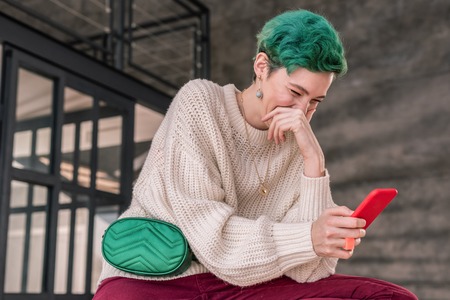 Reading funny story. Green-haired woman laughing out loud after reading funny story on smartphone