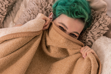 Hiding face. Green-haired cheerful woman hiding her face behind plaid lying in bed in the morningの写真素材