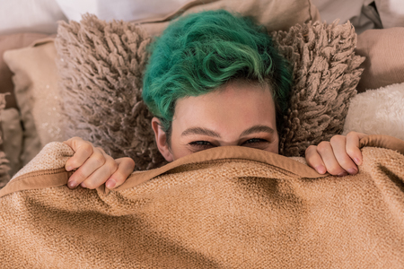 Under plaid. Green-haired cheerful funny woman lying on fluffy brown pillow under plaidの写真素材