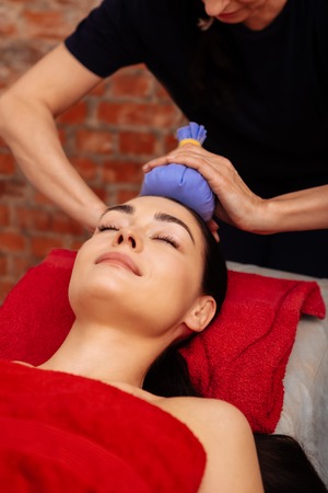 Feeling completely relaxed . Pleasant good-looking woman in red towel having calming session while master attaching herbal bag to her foreheadの写真素材