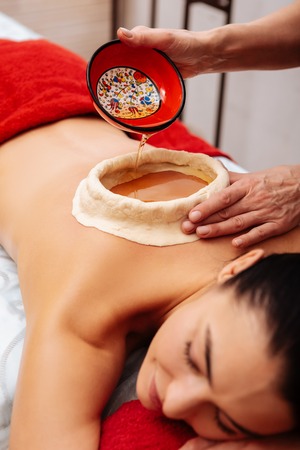 Experiencing Ayurveda treatment. Dark-haired woman having kati vasti procedure in SPA salon while master filling construction with scented oilの写真素材
