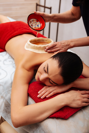 Master filling construction. Relaxed long-haired woman resting on massage bed while master adding oil in special reservoir on backの写真素材