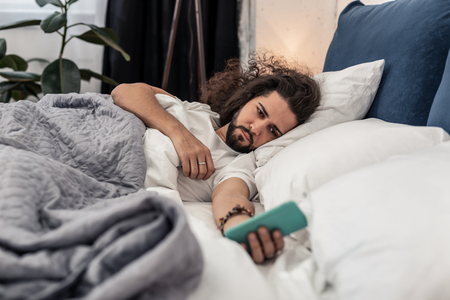 What time is it. Cheerless sleepy man holding his smartphone while checking the time in the morningの写真素材