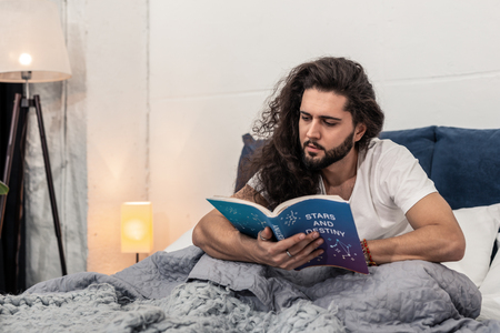 So interesting. Smart long haired man sitting on the bed while being engaged in readingの写真素材