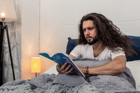 Study of stars. Serious young man sitting on the bed while reading a book on astrologyの写真素材