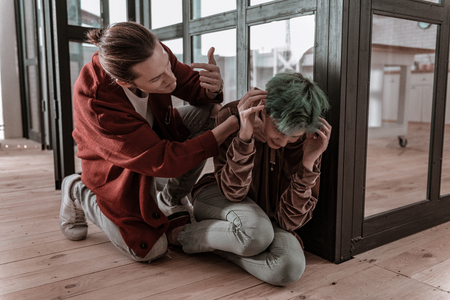 Near glass door. Green-haired woman sitting on floor near glass door suffering from violence of her angry husbandの写真素材