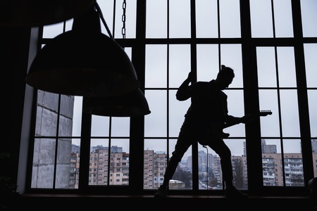 No light. Young rock musician standing near the window with no light and playing musicの写真素材