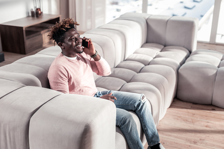 Be positive. Joyful stylish dark-skinned man talking per telephone and sitting in cozy living roomの写真素材