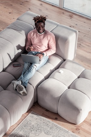 Concentrate on task. Cheerful young man holding laptop on knees while chatting onlineの写真素材