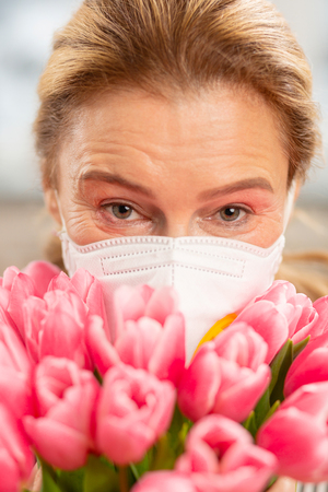 Woman in mask. Mature pleasant blond-haired woman wearing mask and holding pink tulipsの写真素材