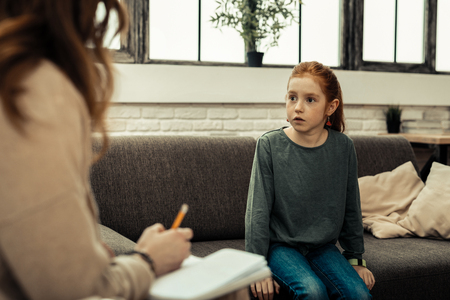 Childish fears. Nice worried girl sitting on the table while looking at the psychologistの写真素材