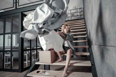 Sitting on stairs. Lonely emotional woman sitting on stairs at home and throwing clothes of ex husbandの写真素材