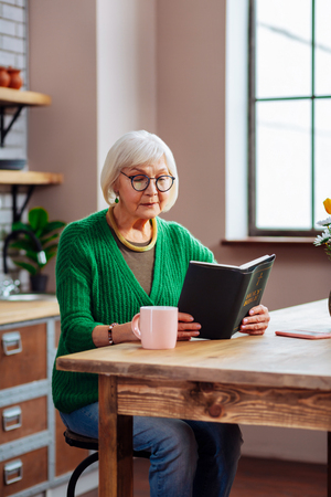 Sitting at kitchen table with Bible. Septuagenarian white-haired dame in stylish glasses and jade green cardigan sitting at kitchen table with Bible in handsの写真素材