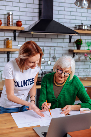 Helping with papers. Appealing focused grey-haired elderly woman wearing green knitted clothes asking young-adult short-haired slim nice lady with volunteer sign t-shirt for helping with papersの写真素材