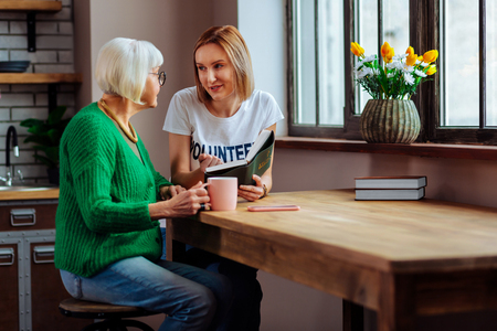 Discussing Bible statements. Charming alluring young fair-haired woman discussing with stylishly worn elegant short-haired pensioner Bible book at kitchen tableの写真素材