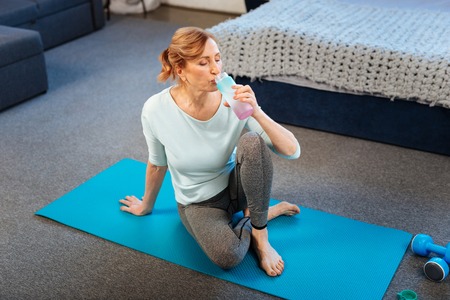 Comfortable clothes. Peaceful fit lady in leggings resting on sportive mat and drinking fresh water after intense trainingの写真素材