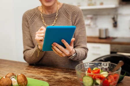 Checking media. Mature lady in beige warm sweater carrying blue tablet and typing on it
