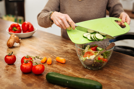 Adding chopped cucumbers. Attentive woman in beige sweater throwing vegetables into glass bowl while having light dinnerの写真素材