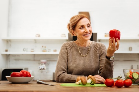 During cooking process. Contented light-haired woman with wide smile lovely looking on pepper in her handの写真素材