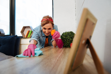 Wooden table. Concentrated light-haired mature woman wearing pink rubber gloves while cleaning...
