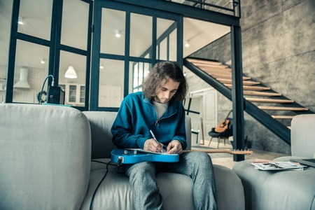 Professional young musician. Focused dark-haired guy writing down notes in special album while...