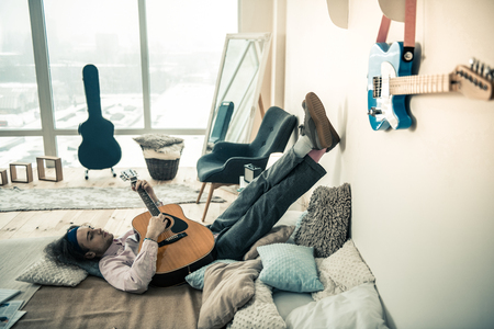Lying upside down. Melancholic young artist leaning on clear wall with his legs while hugging acoustic guitarの写真素材