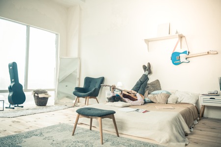 Performing sounds. Good-looking artist lying on bed in bright stylish bedroom while being alone at homeの写真素材
