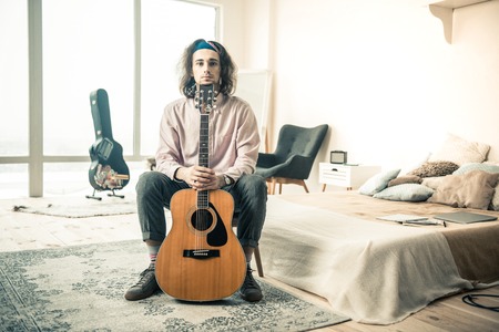 Connected hands. Long-haired young musician resting in stylish bedroom and leaning on a standing guitarの写真素材