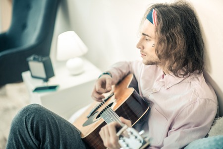 Entertaining himself. Emotional long-haired artist being concentrated on personal musical instrument while being alone at homeの写真素材