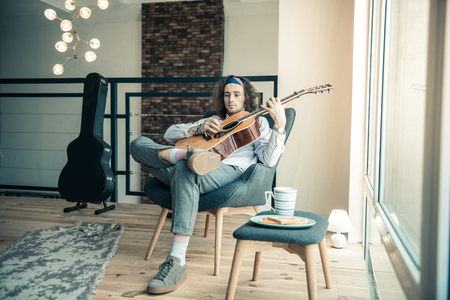 Meditating and thinking. Melancholic young musician relaxing in spacious armchair with trustful acoustic guitarの写真素材