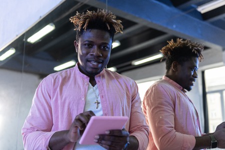 Typing on tablet. Smiling dark-skinned man in light outfit being contented with his tablet while...