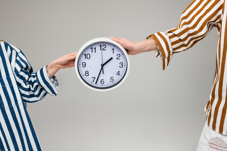 Round wall clock. Women in oversize striped outfits carrying plain wall clock between them with both...