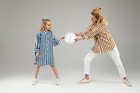 Plain wall clock. Long-haired pretty sisters playfully fighting for round clock while pulling him...