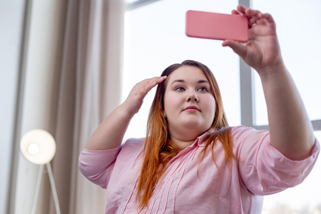 I am beautiful. Pleasant overweight woman fixing her hair while wanting to look pretty on the photoの写真素材