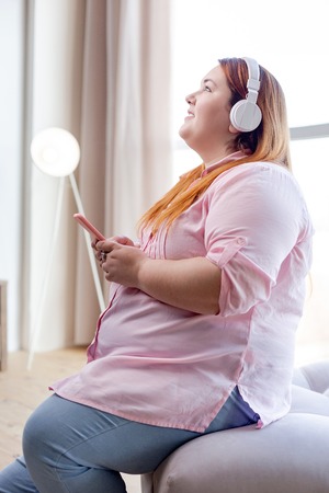 Perfect relaxation. Joyful overweigh woman holding her smartphone while listening to musicの写真素材