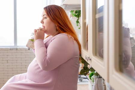 Full of vitamins. Nice plump woman sitting in the kitchen while drinking healthy smoothieの写真素材