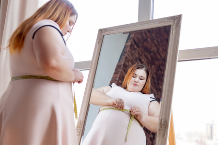 Body measurement. Nice chubby woman measuring her waist while standing in front of the mirrorの写真素材