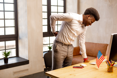 Health problems. Sad young man standing near the table while feeling the pain in his backの写真素材